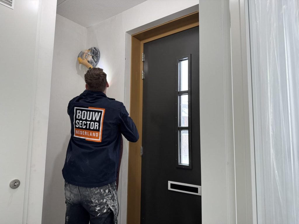 Vakman van Bouwsector Nederland schuurt de wand boven een voordeur met een schuurmachine met afzuiging